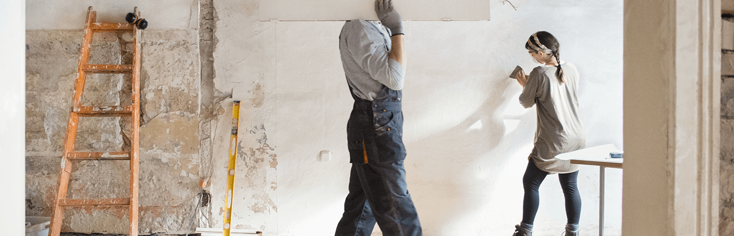 Man carries plaster board whilst women smooths wall during building renovation